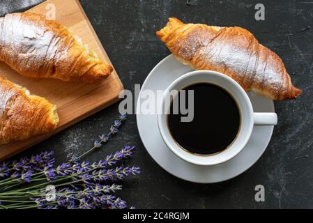 Una tazza di caffè e croissant, mazzo di fiori di lavanda su un tavolo nero, vista dall'alto. Concetto di colazione del mattino Foto Stock