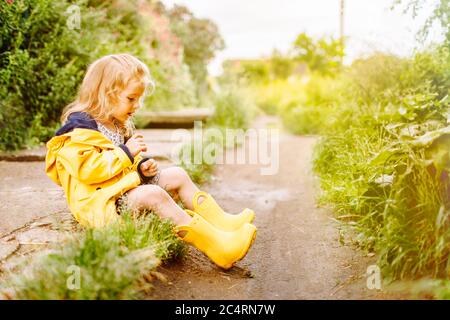 Bambina bionda in impermeabile giallo in piedi e stivali in gomma seduta su una strada dopo una pioggia. Estate, felice infanzia. Foto Stock