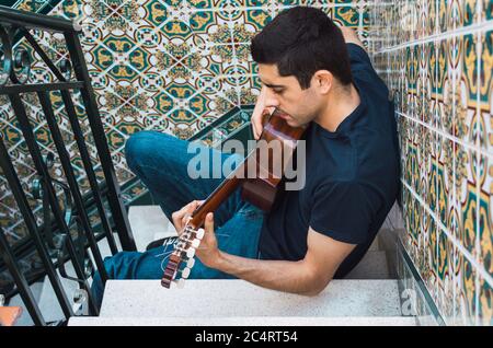Giovane che suona la chitarra seduto su una scala con belle piastrelle. Foto Stock