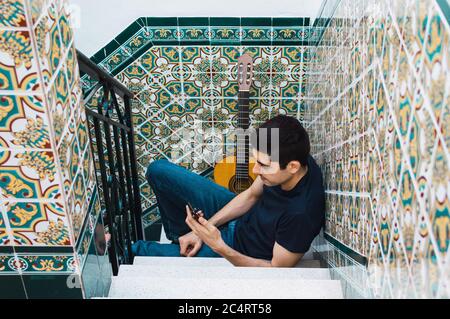 Uomo che usa uno smartphone mentre si riposa dal suonare la chitarra a casa. Foto Stock
