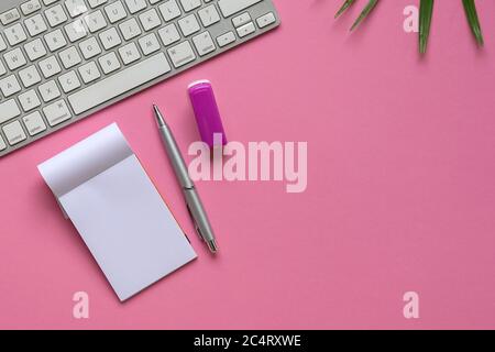 Disposizione piatta di tastiera, cucitrice, blocco note, penna e pianta verde su sfondo rosa. Spazio di copia. Foto Stock