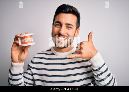 Giovane bell'uomo con barba che tiene denti di plastica dentato su sfondo bianco sorridendo facendo il gesto del telefono con le mani e le dita come parlare sulla Th Foto Stock