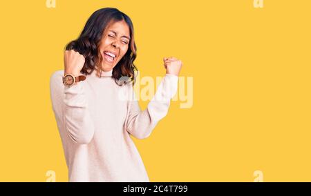 Giovane bella donna mista da corsa con maglione invernale a dolcevita molto felice ed eccitata facendo gesto vincitore con braccia sollevate, sorridenti e urlanti Foto Stock