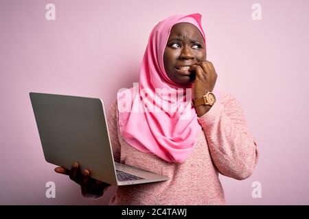 African american più taglia donna che indossa hijab musulmano usando il laptop su sfondo rosa cercando stressato e nervoso con le mani sulla bocca che mordicchiano le unghie. Foto Stock