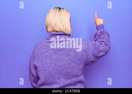 Bella donna bionda Plus taglia indossa un pullover casual turtleneck su sfondo viola che si pone indietro puntando davanti con la mano del dito Foto Stock