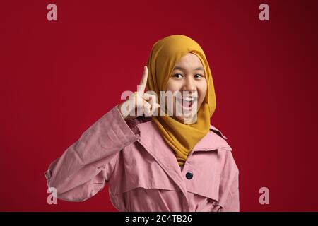 Asian musulmana giovane ragazza adolescente che indossa hijab sorridente per avere una buona idea con il dito rivolto verso l'alto, contro sfondo rosso Foto Stock