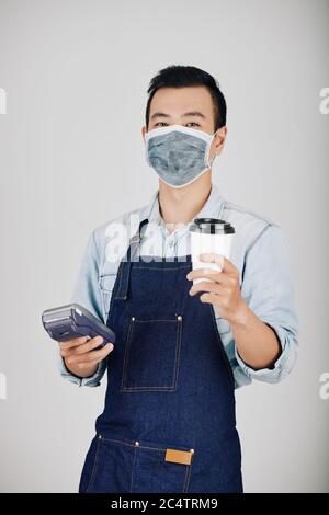 Ritratto del giovane proprietario di una caffetteria in maschera medica che tiene la tazza di caffè e il terminale di pagamento Foto Stock