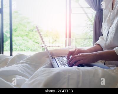 Le mani della donna che scrivono sul computer portatile sul letto all'interno della casa con luce solare dall'esterno attraverso la porta di vetro. Lavorare a casa con relax. Foto Stock