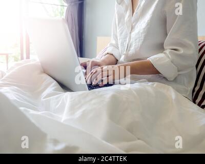 Le mani della donna che scrivono sul computer portatile sul letto all'interno della casa con luce solare dall'esterno attraverso la porta di vetro. Lavorare a casa con relax. Foto Stock