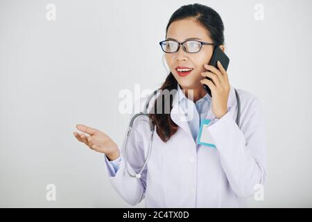 Eccitato giovane medico asiatico generico in occhiali che parla al telefono con un collega o paziente Foto Stock