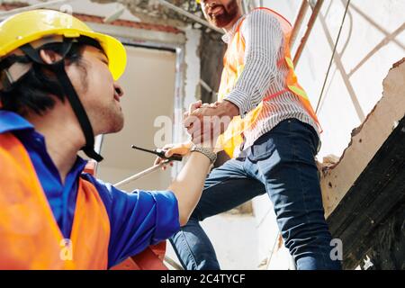 Sorridente lavoratore di costruzione che si stende la mano quando aiutare il collega ad entrare in scaletta accanto a lui Foto Stock