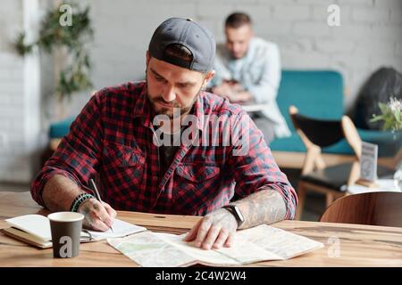 Uomo giovane e bearded messo a fuoco con tatuaggi seduti a tavola di legno in caffè e prendere appunti sul prossimo viaggio in diario Foto Stock