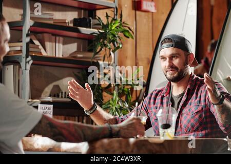 Soddisfare l'uomo bearded giovane che mostra la taglia con le mani ad un amico mentre discutono il surf in caffè estivo Foto Stock