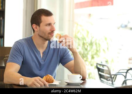 Un uomo pensivo che fa colazione con una tazza di caffè e un croissant sembra gettare la finestra seduta in una caffetteria Foto Stock