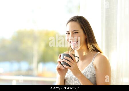 Donna felice a casa tenendo una tazza di caffè si guarda accanto a una finestra in una giornata di sole Foto Stock