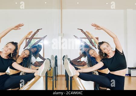 Ballerini giovani sorridenti graziosi che mettono i piedi su barre quando fanno l'esercitazione di stiramento per le gambe Foto Stock