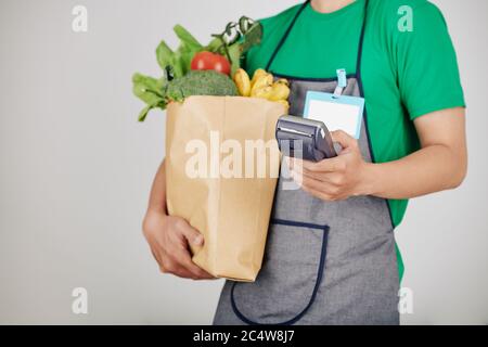 Immagine ritagliata di un supermarket operaio che trasporta il pacchetto di alimentari freschi e di terminale di pagamento Foto Stock