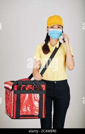 Vietnamita giovane donna corriere con grande sacchetto di cibo che indossa maschera medica quando si parla al telefono e consegna ordini Foto Stock