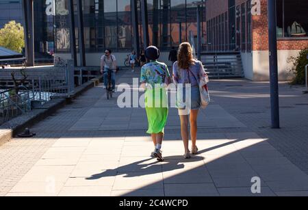 Due ragazze che camminano fianco a fianco sulla strada pedonale in bei vestiti colorati visti da dietro. Uno indossando lungo abito verde e gli altri pantaloni caldi je Foto Stock