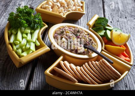 Insalata di sottaceti per tonni servita con carote e bastoncini di sedano, cetrioli freschi, cracker e scorze di maiale in ciotole su un rustico tavolo di legno, Foto Stock