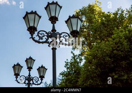 Lampione con una tripla lanterna con motivi e inserto in vetro nella vista del parco dal basso sullo sfondo di alberi e cielo. Foto Stock