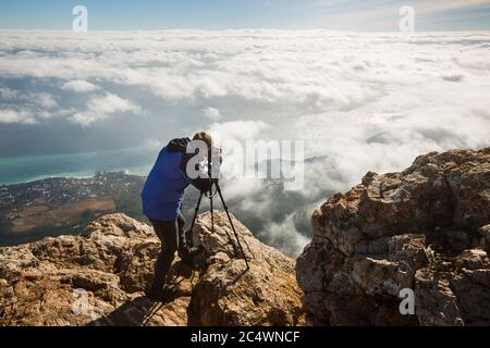 Uomo in piedi con un treppiede e una fotocamera su un'alta montagna cima sopra le nuvole, la città e il mare. Fotografo professionista che regola le impostazioni dslr su roccioso Foto Stock