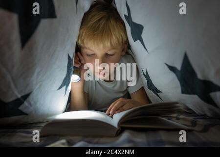 Un ragazzino legge un libro con una torcia sotto le coperture di notte. Foto Stock