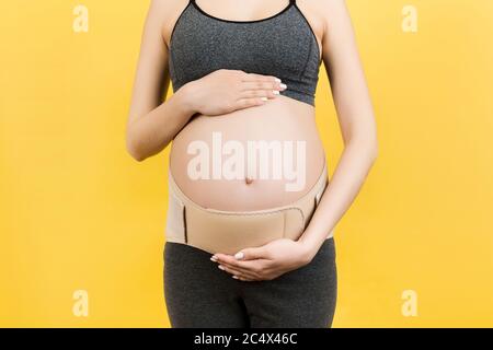 Primo piano di donna incinta in elastico gravidanza bendaggio per fare il dolore andare via a sfondo giallo con spazio di copia. Supporto addominale ortopedico BE Foto Stock