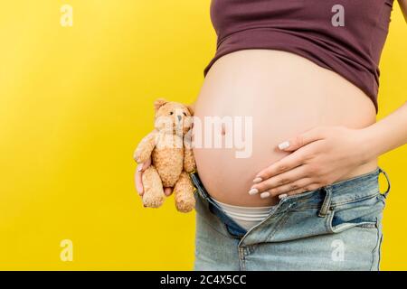 Primo piano di donna incinta in jeans unzipped abbracciando orsacchiotto vicino al ventre a sfondo colorato con spazio di copia. Aspettandosi un concetto del bambino. Foto Stock