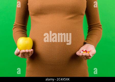 Primo piano di donna incinta che fa una scelta tra una mela e un mucchio di pillole a sfondo colorato con spazio di copia. Assunzione di vitamine durante il gravidanza Foto Stock