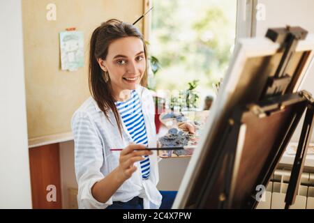 Bella ragazza sorridente in camicia bianca e T-shirt a righe tenendo in mano pennello guardando felice in macchina fotografica mentre disegnando sul cavalletto a casa Foto Stock