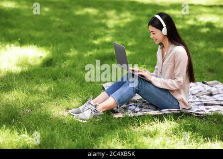 Sorridente ragazza asiatica che si rilassa con laptop e cuffie sul prato nel parco Foto Stock