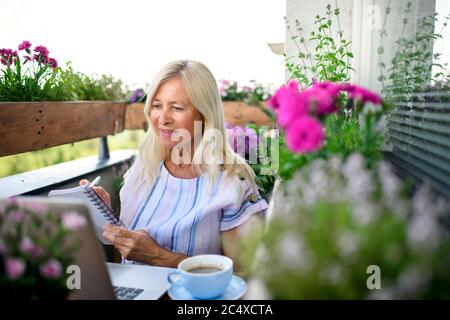 Donna anziana attiva con computer portatile che lavora sul balcone, ufficio domestico. Foto Stock
