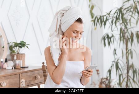 Ragazza felice che controlla sui social media mentre si applica la crema Foto Stock