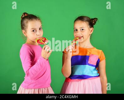 Sorelle con lollipop rotondi e lunghi. Ragazze con volti felici posa con caramelle su sfondo verde. Trattamento e concetto di dolci. I bambini mangiano i grandi caramelle dolci colorate. Foto Stock