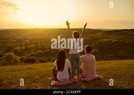Vista posteriore di una famiglia felice riposarsi guardando un bellissimo tramonto in serata sulla natura. Foto Stock
