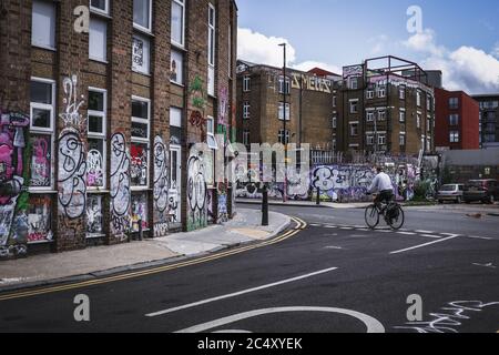 L'uomo fa una bicicletta passando i graffiti su un edificio derelict a Hackney Wick a East London, durante il blocco covid-19 Foto Stock