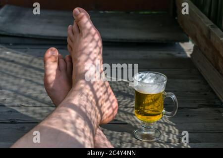 Piedi di un uomo che riposa con una tazza di birra leggera su un pavimento di legno Foto Stock