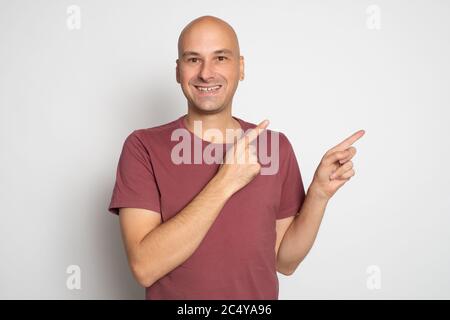 Ritratto di un uomo calvo felice in una t-shirt rossa che punta le dita via allo spazio di copia isolato su sfondo grigio Foto Stock