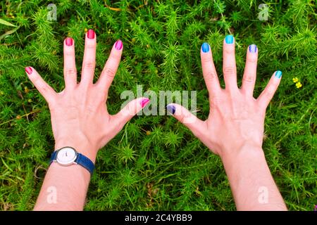 Le mani della donna con ogni chiodo di un colore diverso che indossa un orologio su uno sfondo vegetale Foto Stock