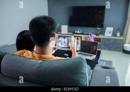Asian bella donna incinta e marito che guarda ecografia su un divano nel soggiorno a casa. Concetto di assistenza sanitaria di gravidanza. Foto Stock