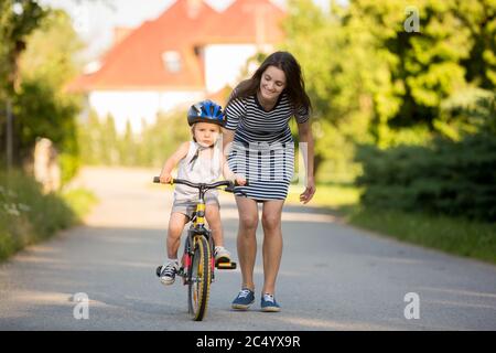 Giovane madre, aiutante suo figlio ad imparare a guidare una bicicletta, tenendolo e insegnandolo a andare in bicicletta Foto Stock