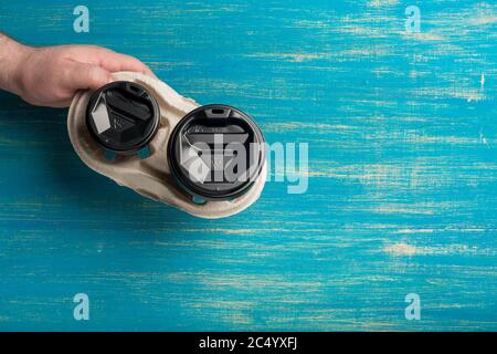 Due tazze di carta monouso di caffè e un portabicchieri in mano maschile su sfondo di legno blu. Foto Stock