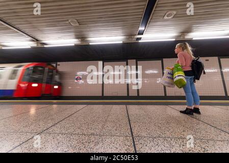 Giovane donna adulta con borse per lo shopping che aspettano sulla piattaforma della metropolitana di Londra con il treno della metropolitana che arriva alla stazione. Lone donna in attesa Foto Stock