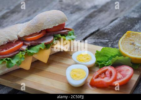 Un appetitoso panino con prosciutto, formaggio, pomodori e lattuga giace su un tavolo con un uovo e limone, su un tavolo di legno, stile artigianale Foto Stock