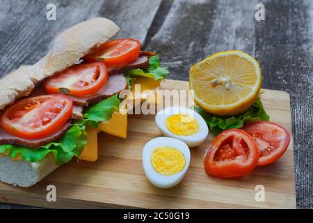 Un appetitoso panino con prosciutto, formaggio, pomodori e lattuga giace su un tavolo con un uovo e limone, su un tavolo di legno, stile artigianale Foto Stock