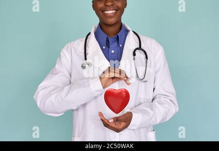 Sorridente donna africana dottore indossare camice bianco tenendo il cuore rosso nelle mani. Foto Stock