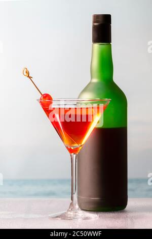 Cocktail fatto in casa dall'aspetto delizioso con ciliegie in un bicchiere di martini davanti a una bottiglia di vino su uno sfondo fuori fuoco. Vacanza e tempo libero conc Foto Stock