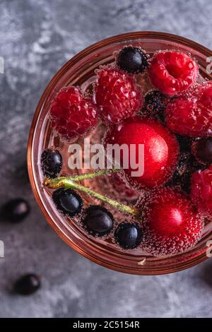 Bevanda fresca di acqua frizzante fredda con ciliegia, lampone e ribes in vetro rosso sfaccettato su fondo di pietra in cemento, bevanda estiva di dieta, t Foto Stock