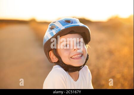 Ritratto di sorridenti grazioso piccolo bambino caucasico in blu sport casco e camicia bianca sul tramonto estivo all'aperto Foto Stock